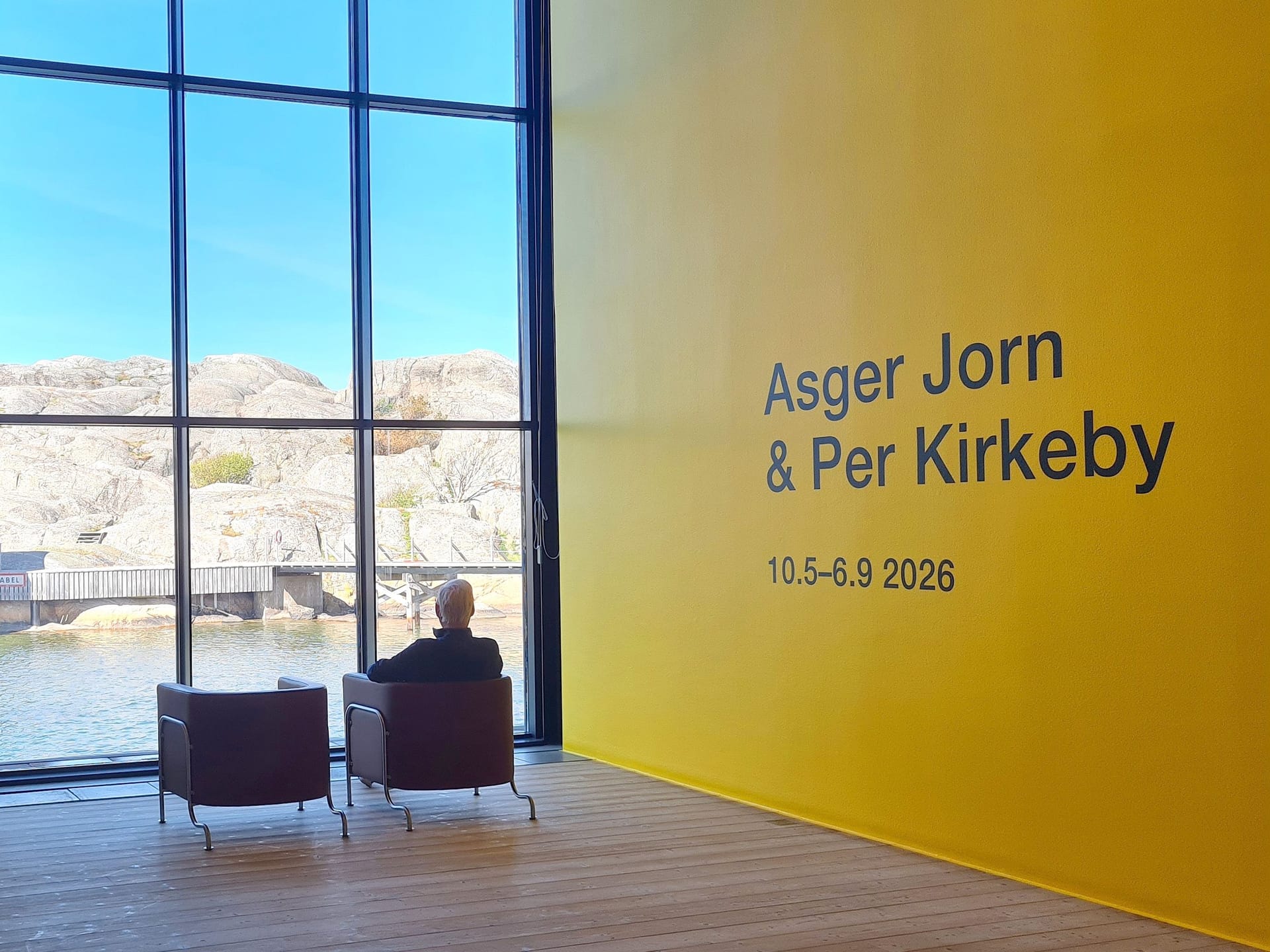 Person i fåtölj tittar på utsikten genom ett stort fönster, bredvid texten "Asger Jorn & Per Kirkeby"