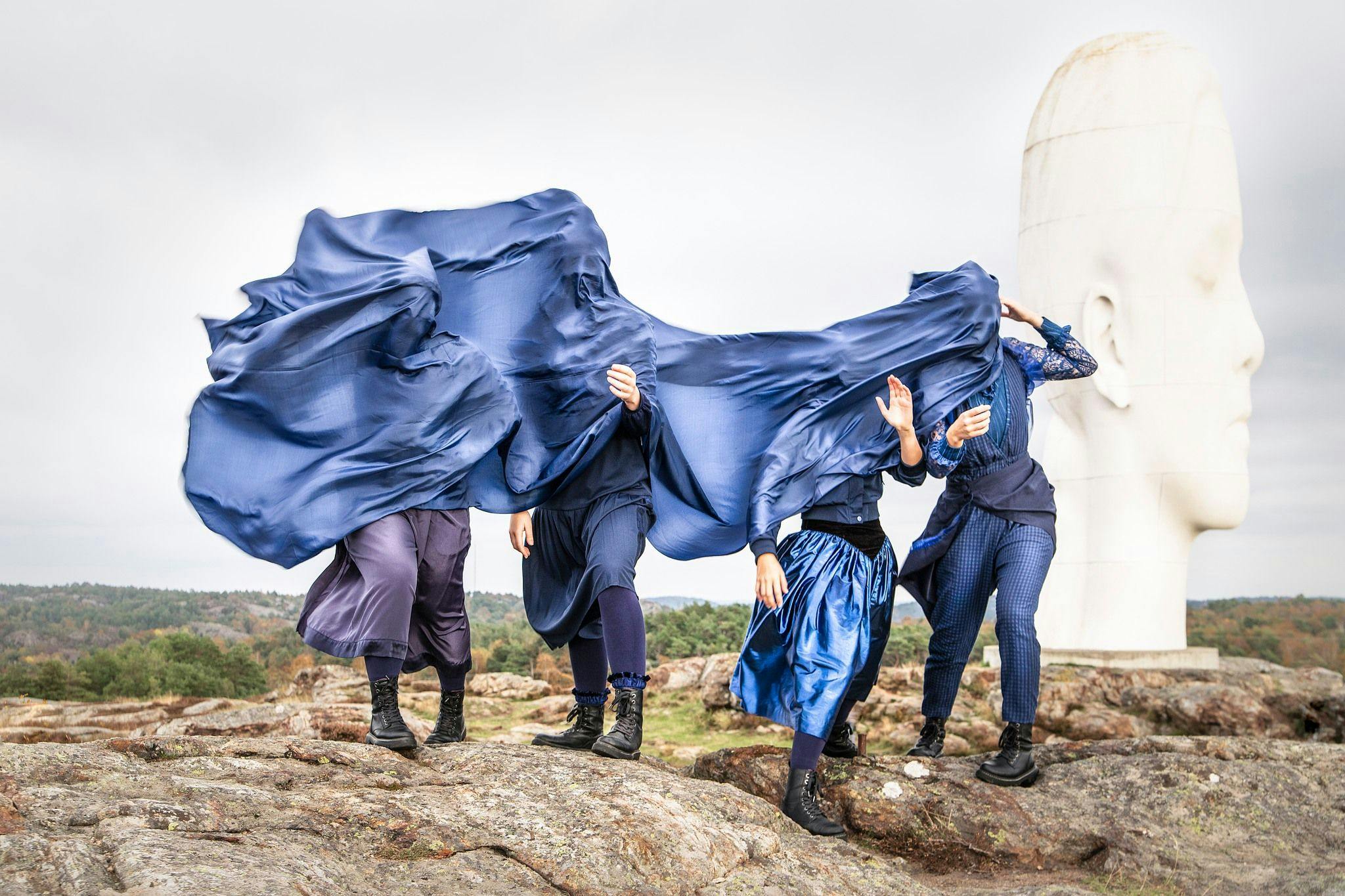 Fyra personer under ett svepande blått tyg. Utomhus, framför en stor skulptur av ett ansikte.