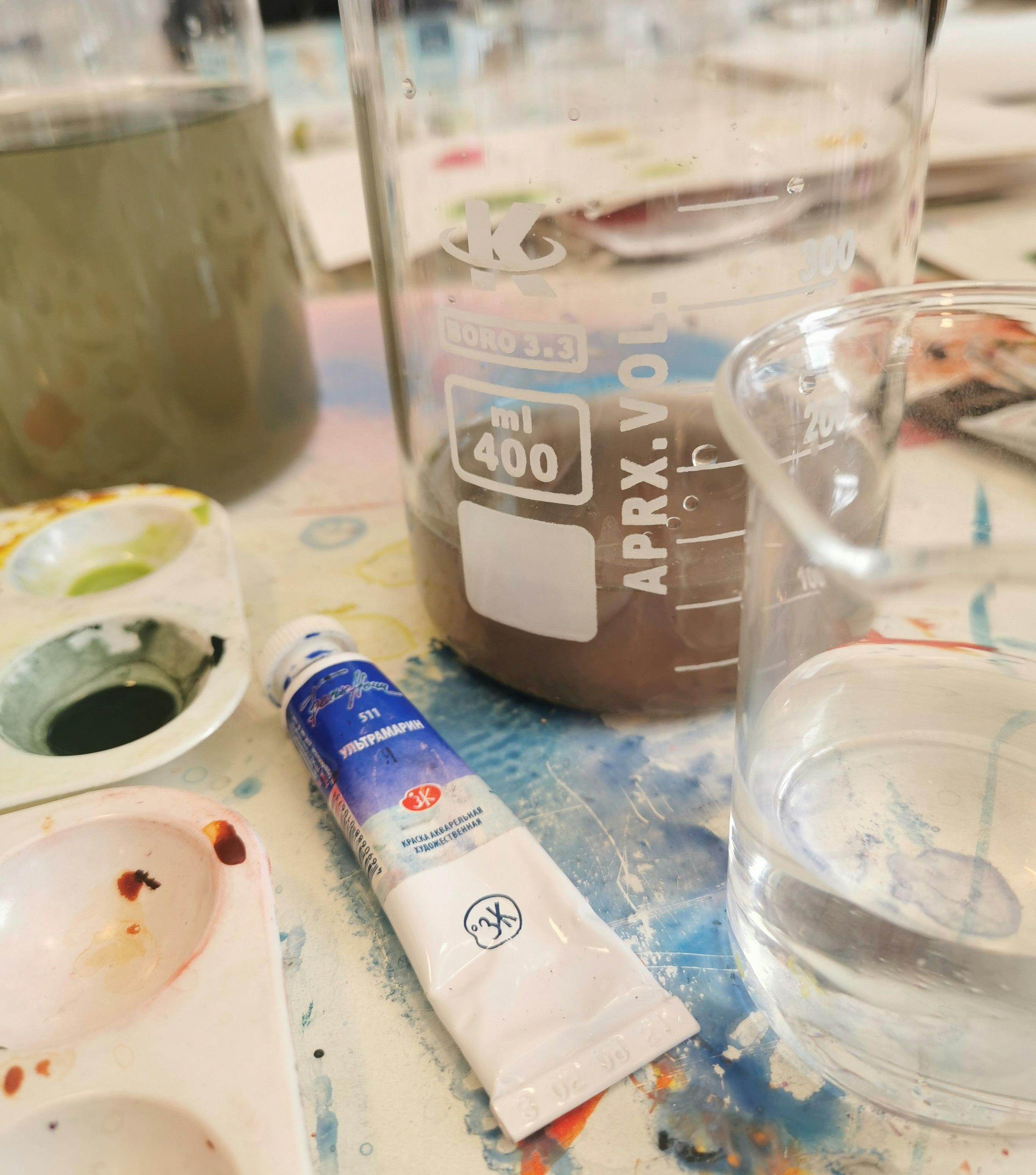 Fotografi av palett med färg, en akvarelltub och ett mätglas med brunfärgat vatten i.