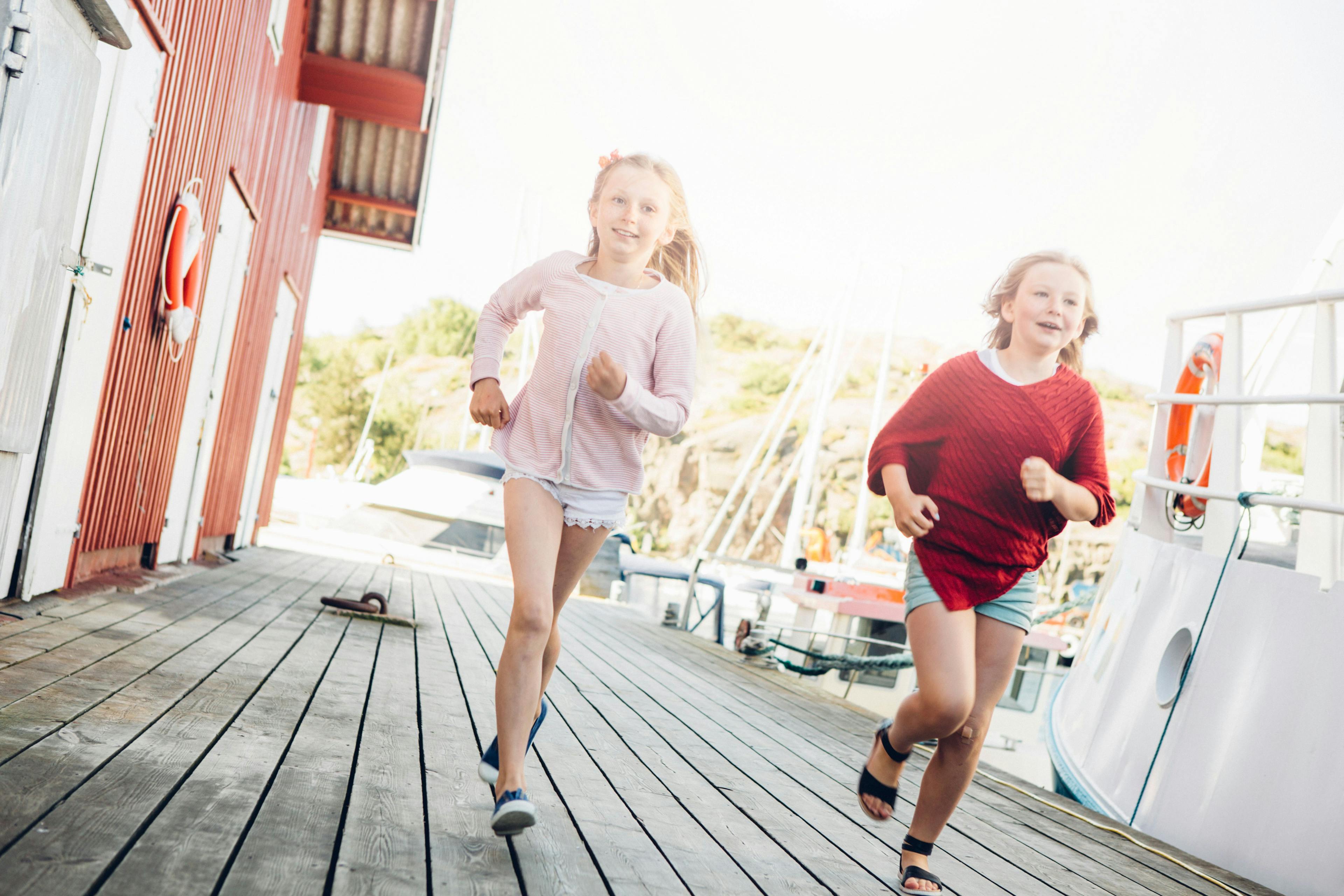 Två barn springer på en brygga bredvid en röd sjöbod, sommartid