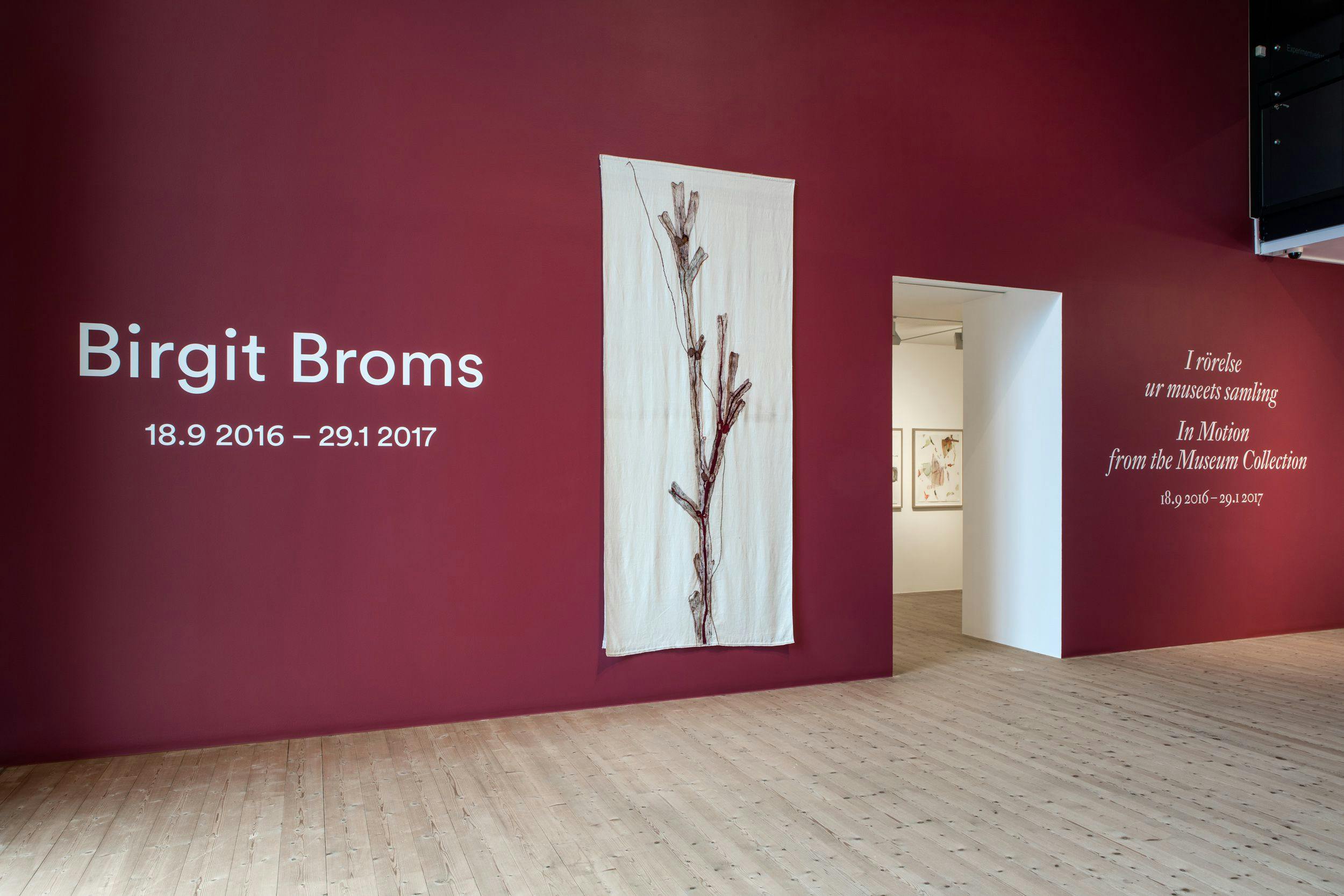 Entrén till utställningen, en vinröd vägg med vit text på, samt ett horisontellt, långsmalt, ljust textilverk.