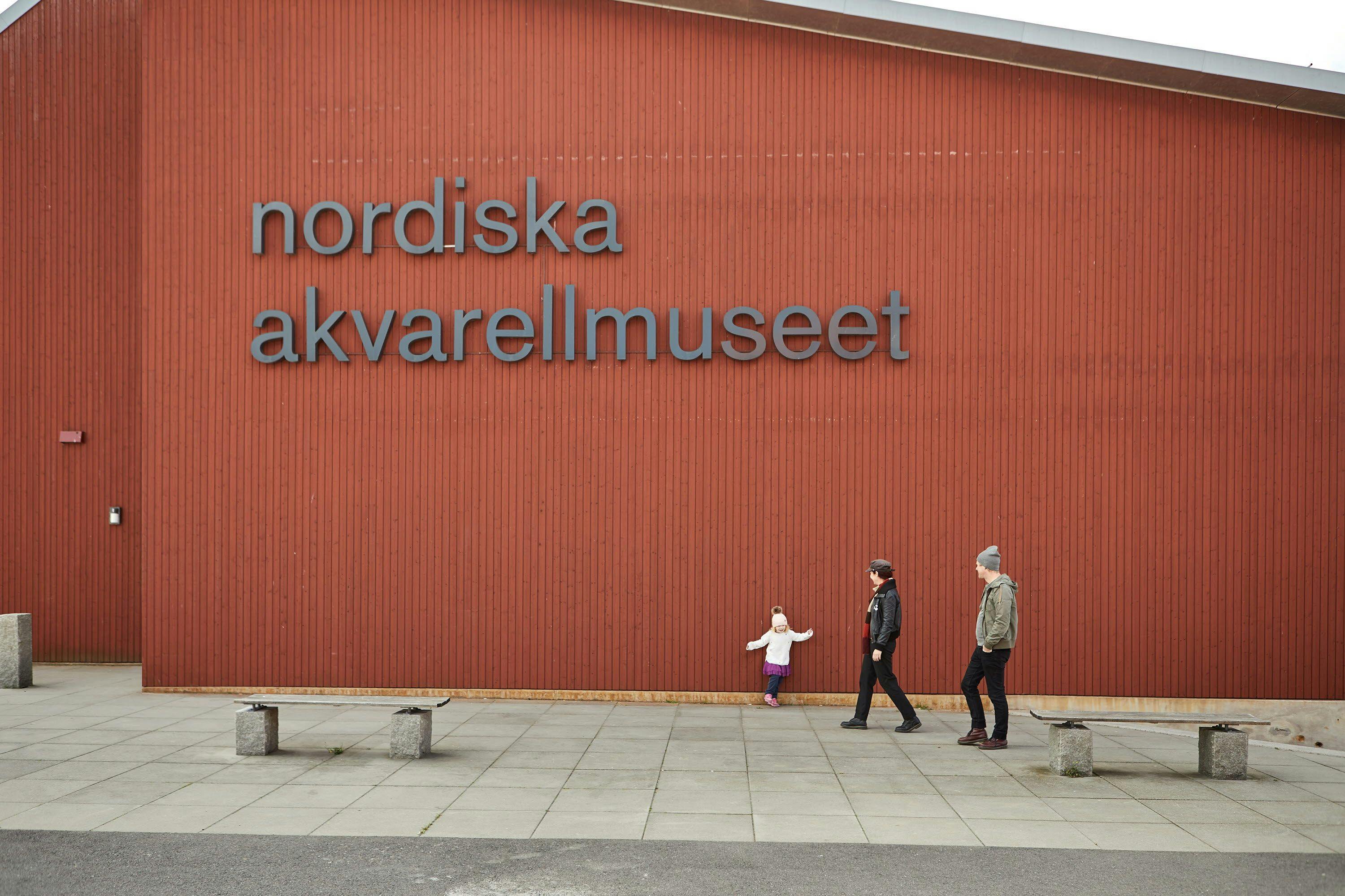 Två vuxna och ett barn går framför en faluröd vägg med texten "Nordiska Akvarellmuseet"