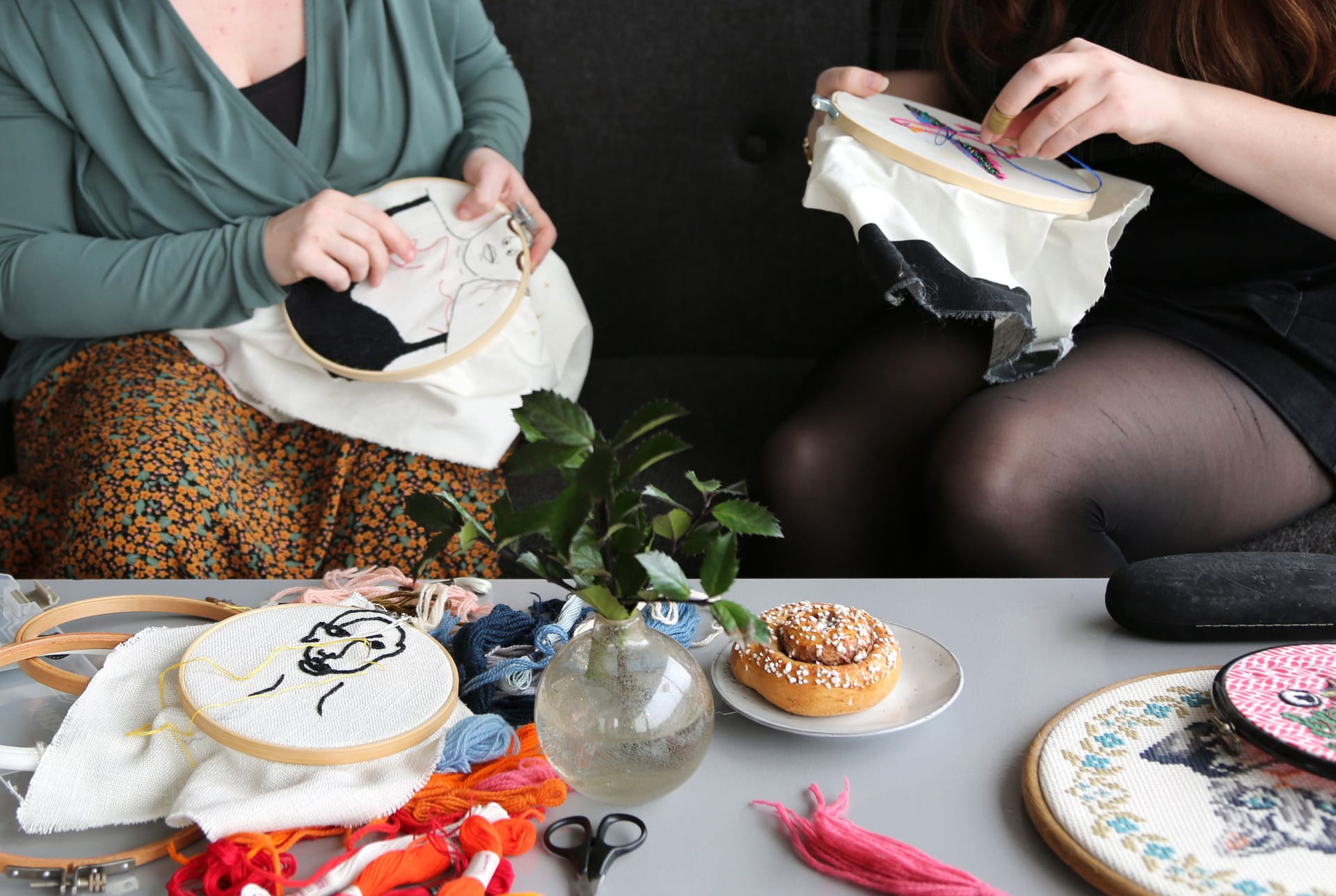 Två personer som broderar, bakom ett bord med textilmaterial och fika