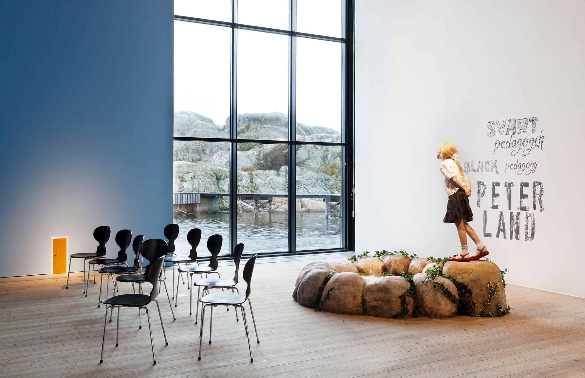 Exhibition room with one blue wall and one white wall. Black chairs and a fountain depicting a child by a pond.