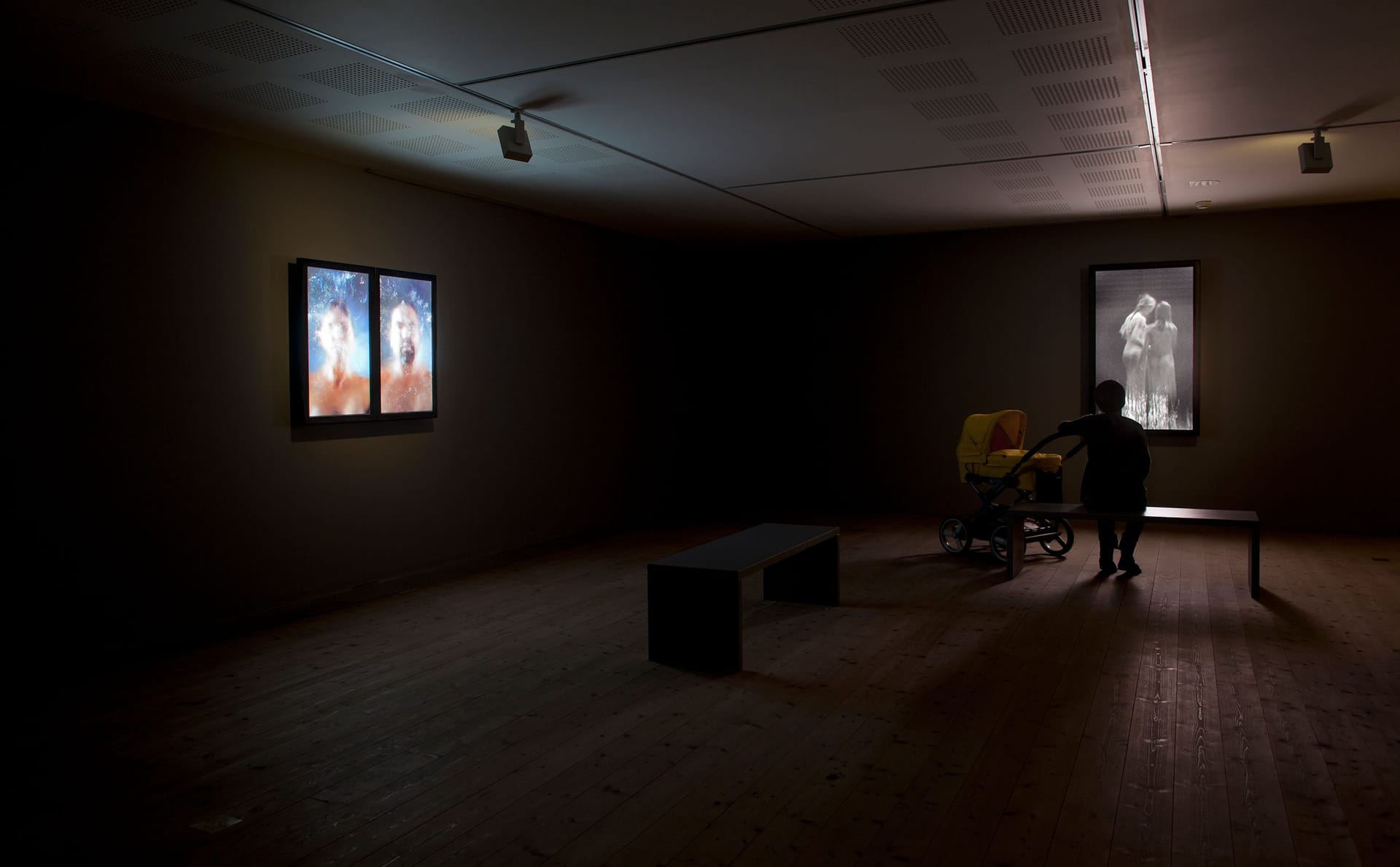 Dimly lit exhibition room with two video works. A visitor with a stroller sits on a bench.