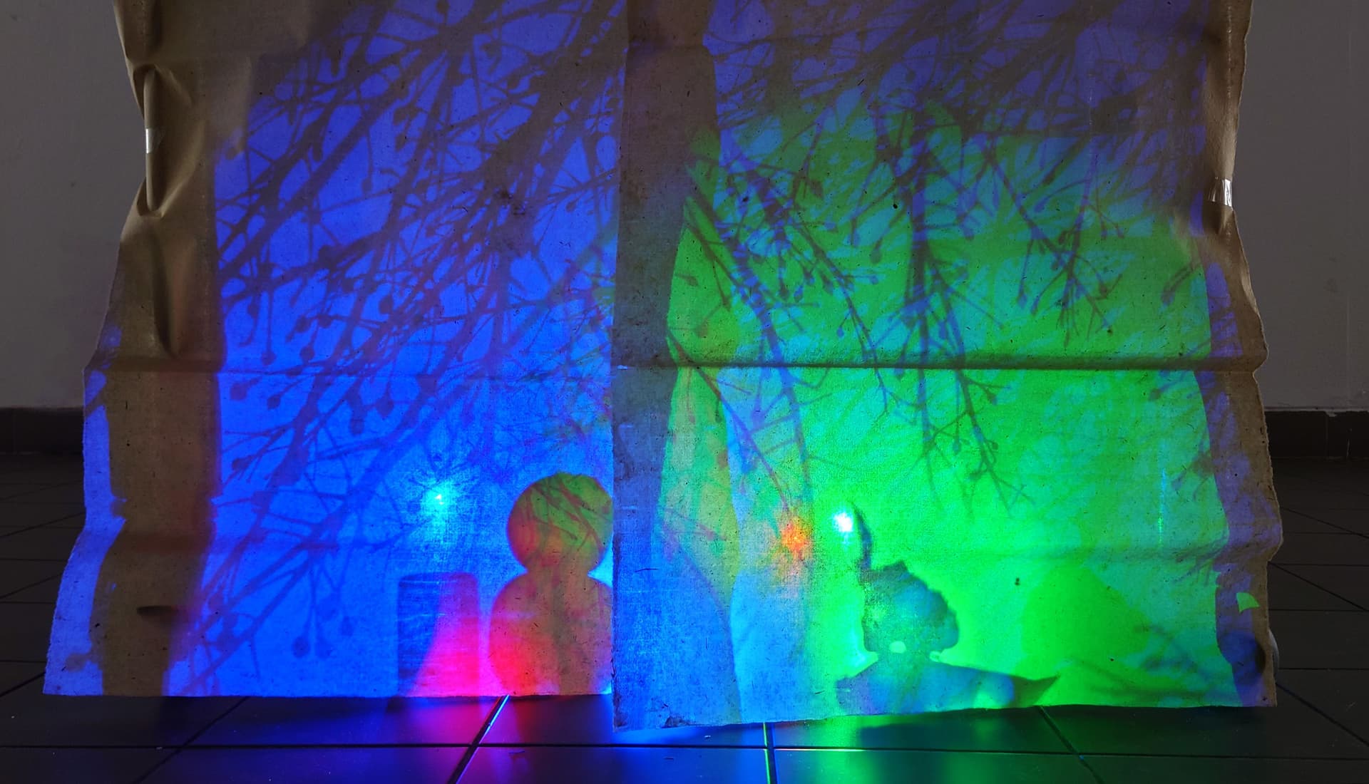 Siluetter av grenar och former bakom ett skynke, ljussatt med blått och grönt ljus