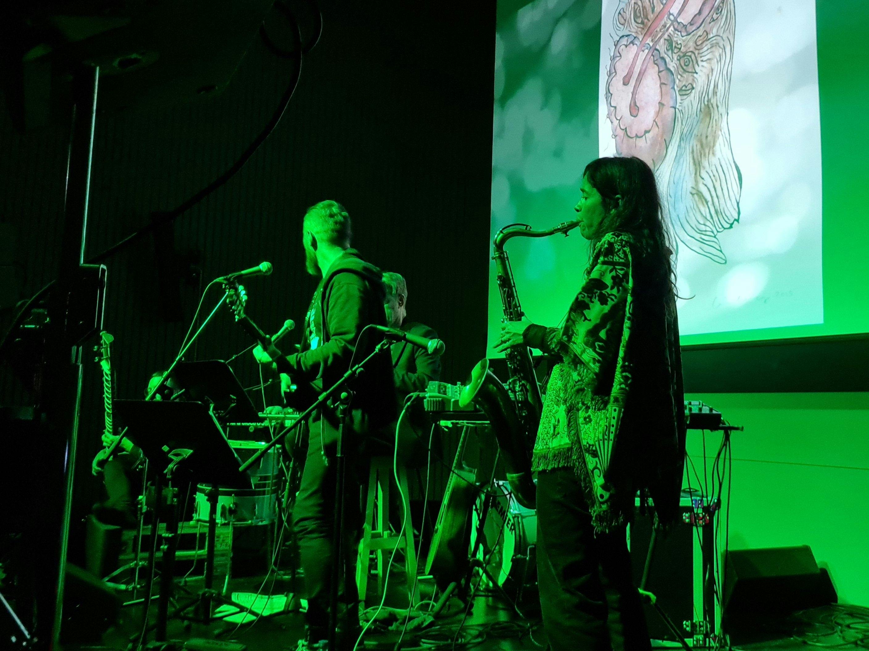 Photo of musicians on stage in green lighting, with saxophonist closest to camera