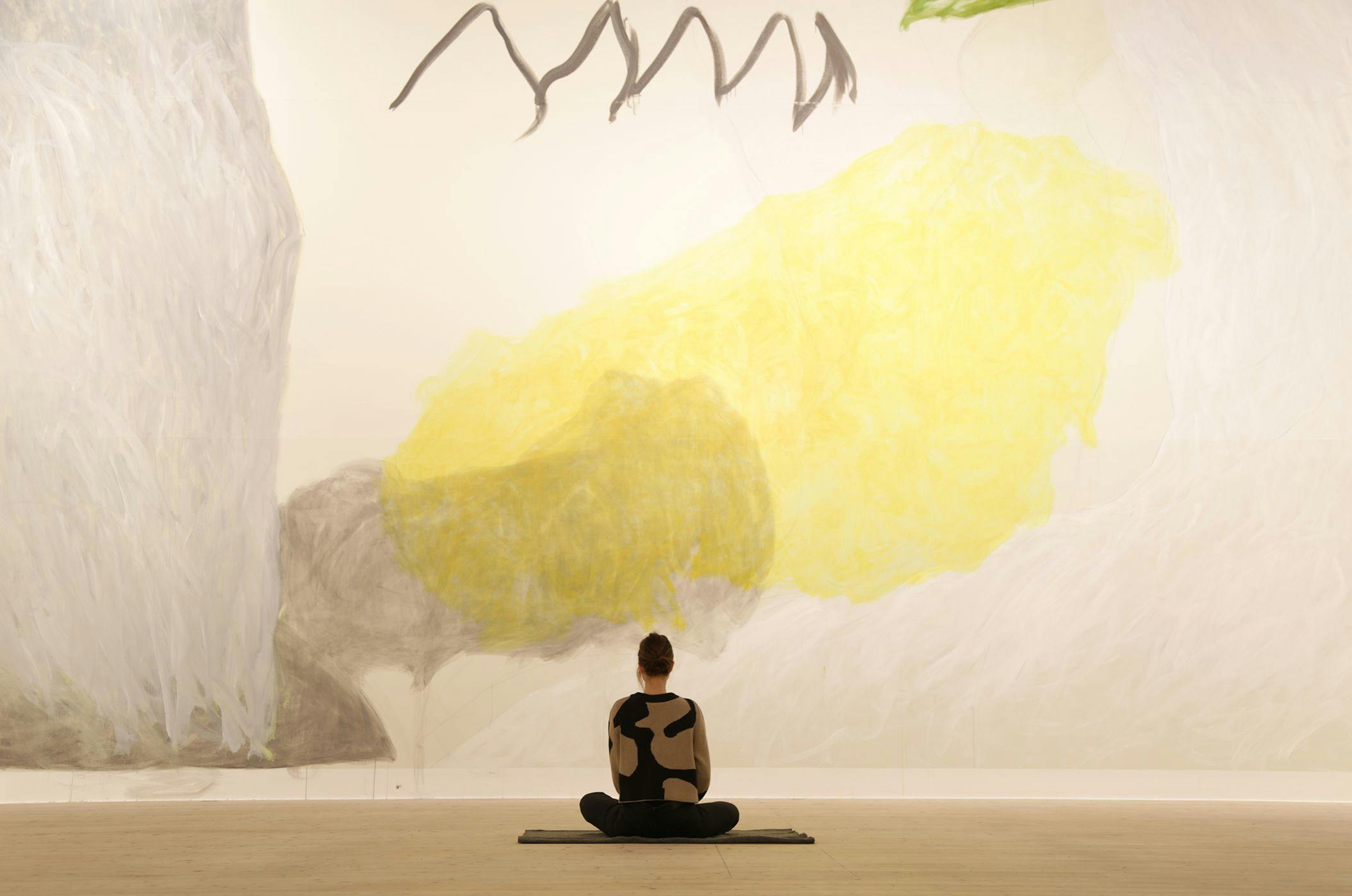 Person sits on the floor with back towards camera, in front of large mural in yellow and gray.