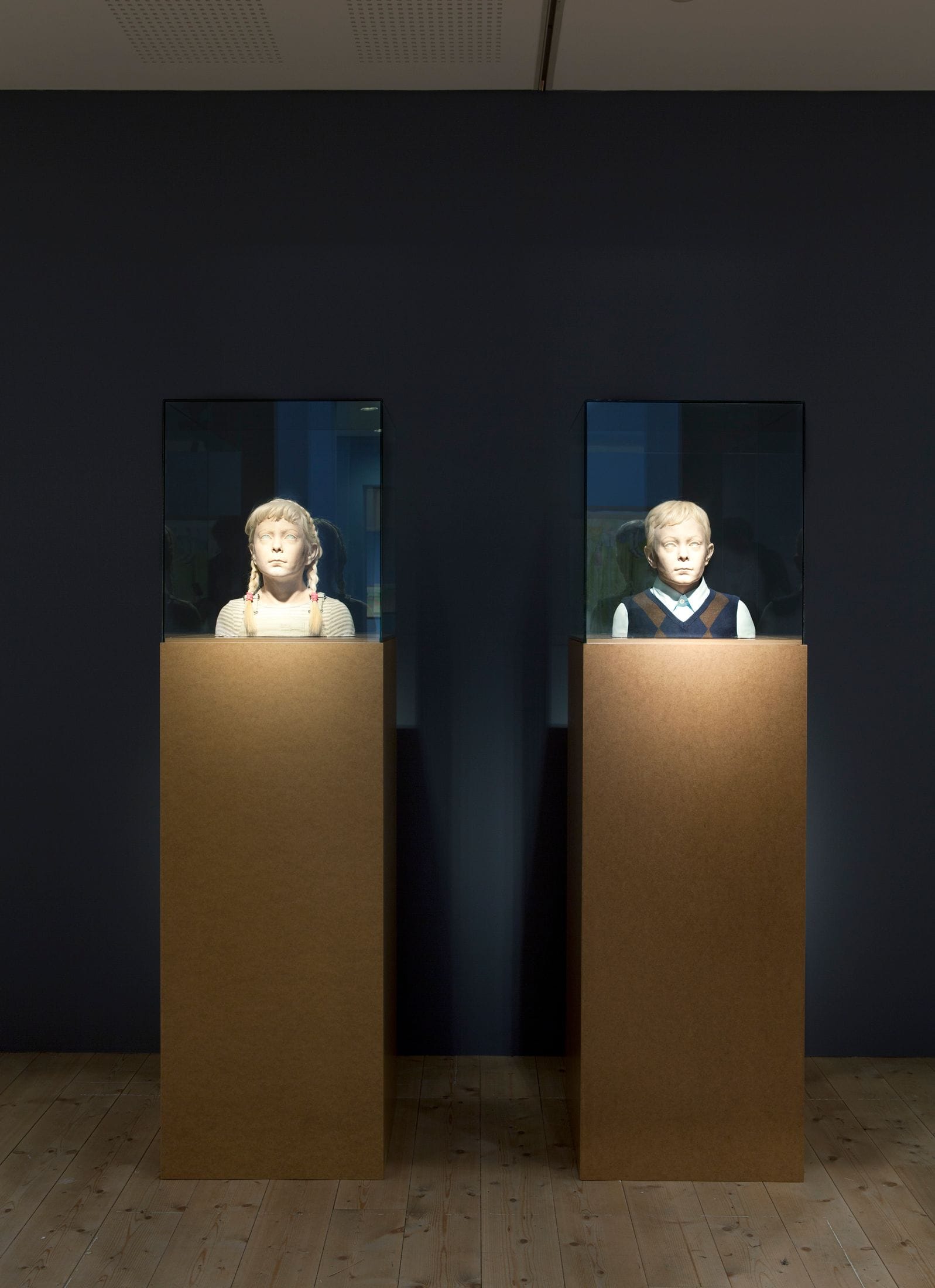 Two sculptures in front of a black wall, depicting busts of children.