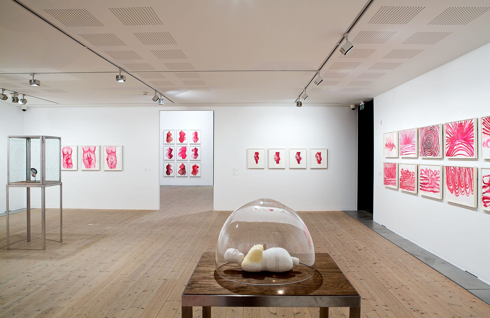 Utställningsrum med röda gouachemålningar av Louise Bourgeois samt skulptur i podium i förgrunden