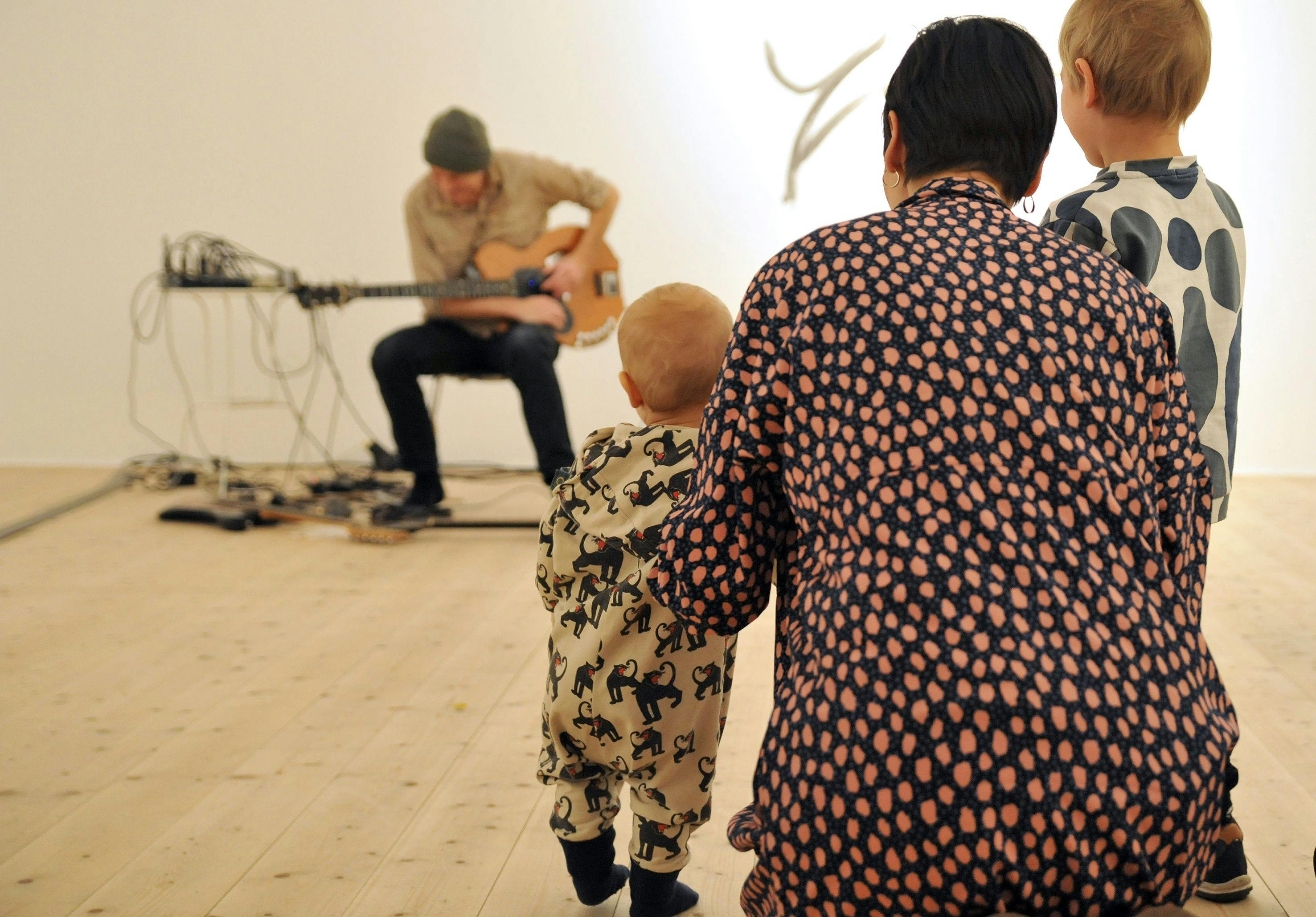 Musiker med gitarr, i förgrunden ryggen på en vuxen och två barn som lyssnar