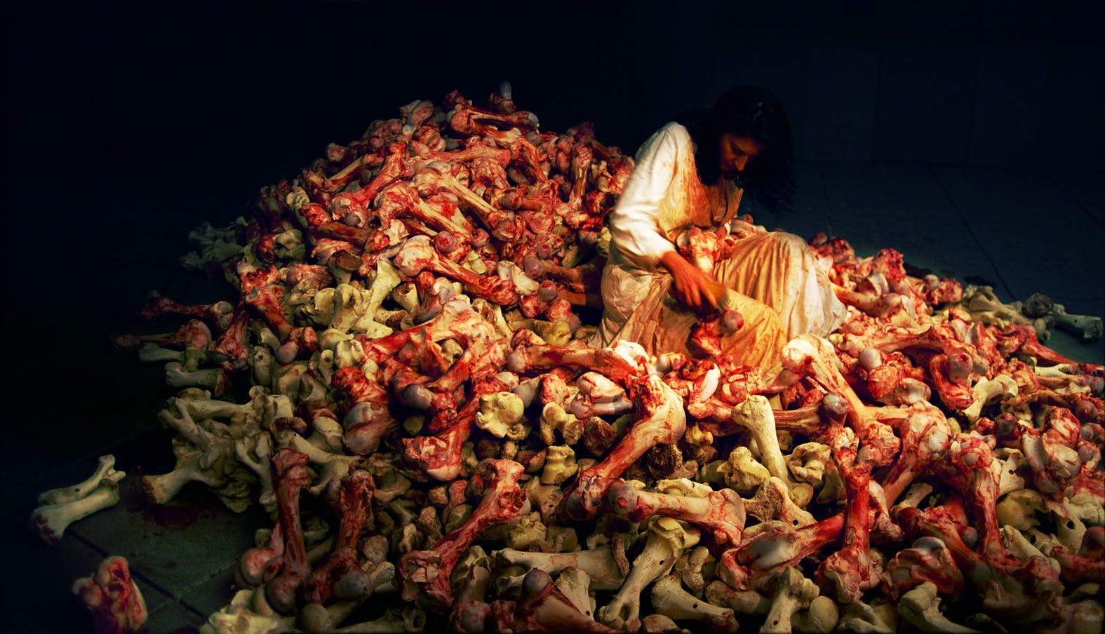 Photo of woman (the artist) sitting on a pile of bloody skeleton bones which she polishes with a coarse brush. From the performance Balkan Baroque by Marina Abramović in 1997